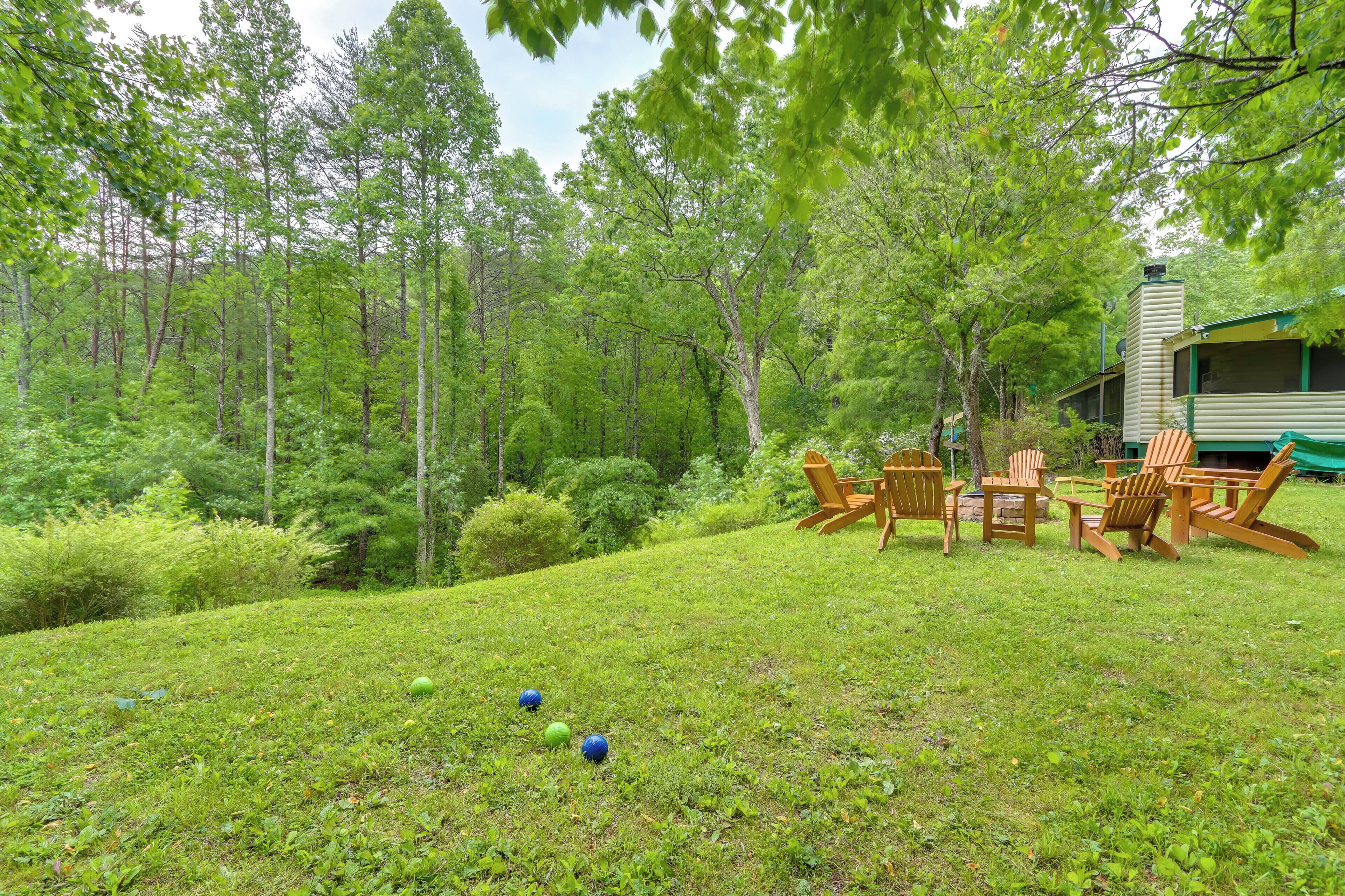A grassy area with Adirondack chairs and a table, surrounded by trees and a cabin.