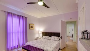 Un dormitorio con cama, una banca, ventilador de techo, mesita de noche y una puerta que da a otra habitación.
