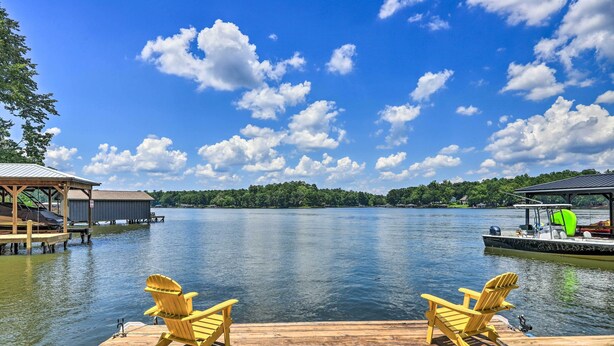 A wooden dock with two Adirondack chairs facing a calm lake, surrounded by docked boats and a boat with a green canopy. - Lakefront Eatonton Getaway w/ Dock & Grill! (Eatonton)