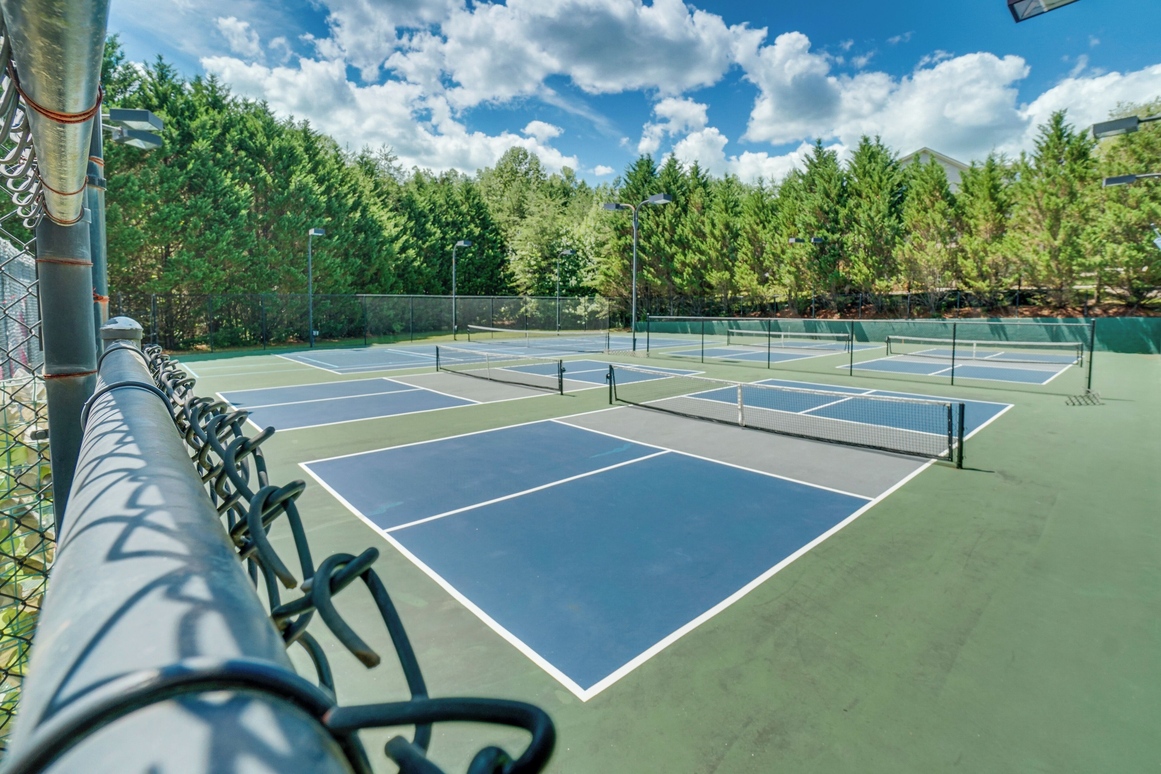 A tennis court with multiple courts, surrounded by a chain-link fence and trees.