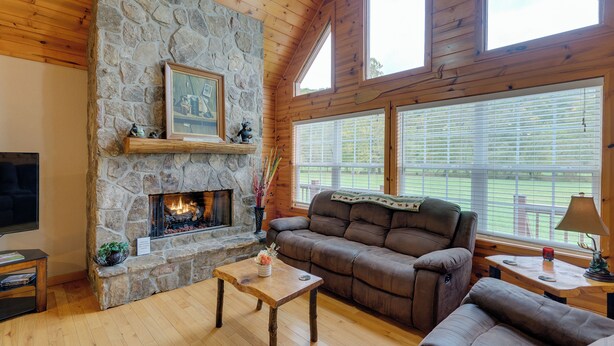 A cozy living room with a stone fireplace, wooden ceiling, and large windows.