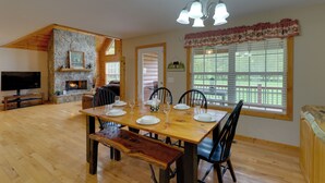 A dining area with a wooden table set for six, a stone fireplace, a television, and a large window with blinds.