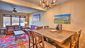 A dining area with a wooden table, chairs, and a chandelier. There is a sofa with a colorful throw blanket and a window with a view of trees. - 7984: Pet-friendly Townhome w/ Grill! (Salida)