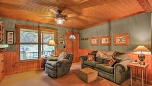 A cozy living room with wooden walls and ceiling, a large window, a ceiling fan, and a fireplace. - Cozy Pine Mountain Cabin w/ Screened Porch & Yard! (Pine Mountain)