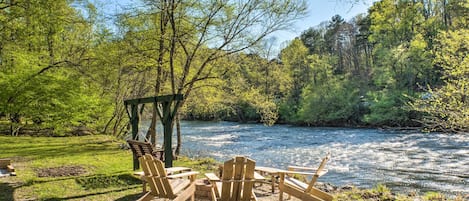 A riverside outdoor seating area with wooden chairs and tables, surrounded by trees and a clear sky.