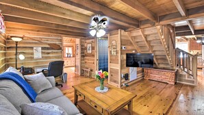 Interior de una cabaña de troncos con techo de madera, un sofá, una mesa de centro con un florero, un escritorio con una lámpara, un televisor y una escalera.