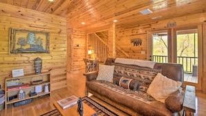 A cozy cabin interior with a wooden interior, a sofa with patterned cushions, a small table with a lamp, and a staircase leading upstairs. - Cozy Hiawassee Cabin w/ Fire Pit & Mtn Views! (Hiawassee)