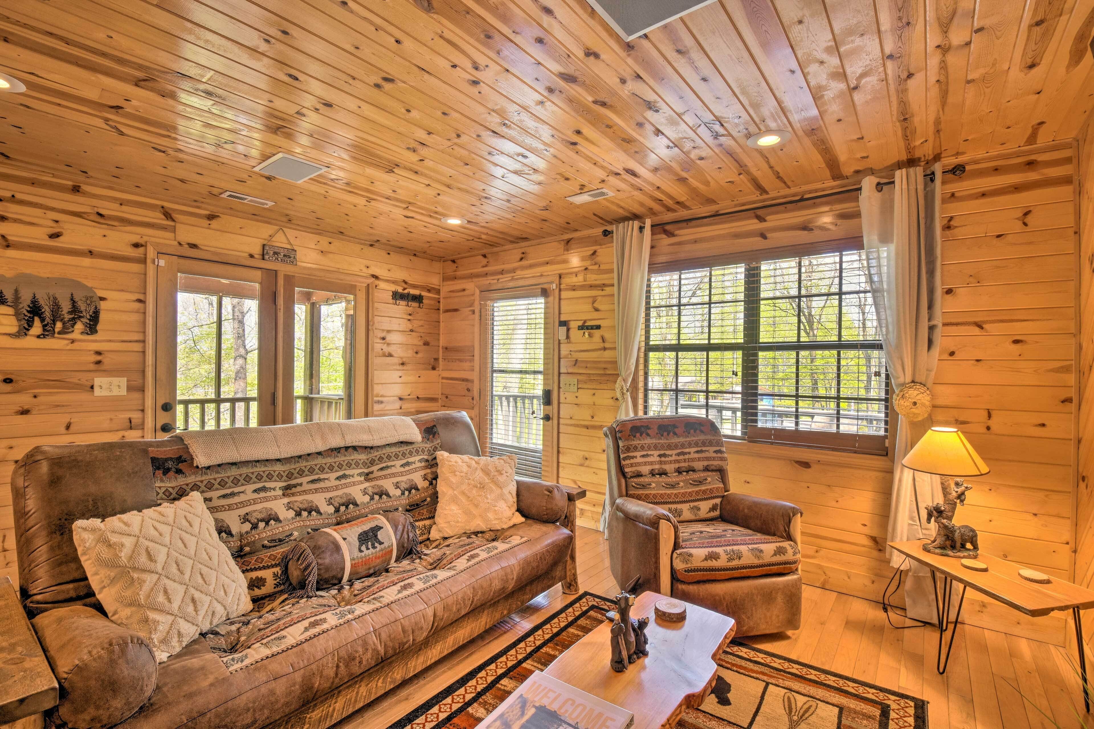 A cozy cabin interior with a wooden ceiling, a sofa, a chair, and a small table with a lamp.