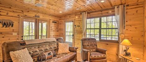 A cozy cabin interior with a wooden ceiling, a sofa, a chair, and a small table with a lamp.