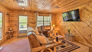 A cozy log cabin interior with a wooden ceiling, a large TV mounted on the wall, a sofa, a chair, and a small table with a lamp. - Cozy Hiawassee Cabin w/ Fire Pit & Mtn Views! (Hiawassee)