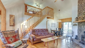 A spacious living area with a wooden staircase, a cozy sofa, a dining table, and a fireplace. - Blairsville Cabin in the Pines 1 Mi to Lake (Blairsville)