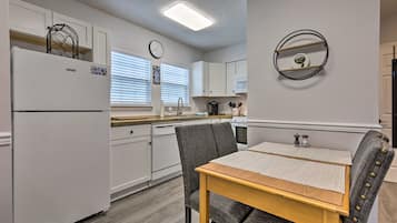 Une cuisine de taille réduite avec des meubles blancs, un réfrigérateur, une table à manger avec des chaises et une horloge fixée au mur.