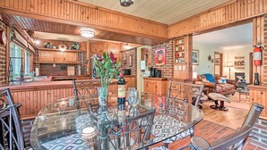 A wooden interior with a dining area featuring a glass table and metal chairs, a kitchen with wooden cabinets, and a living area with a sofa and armchairs.