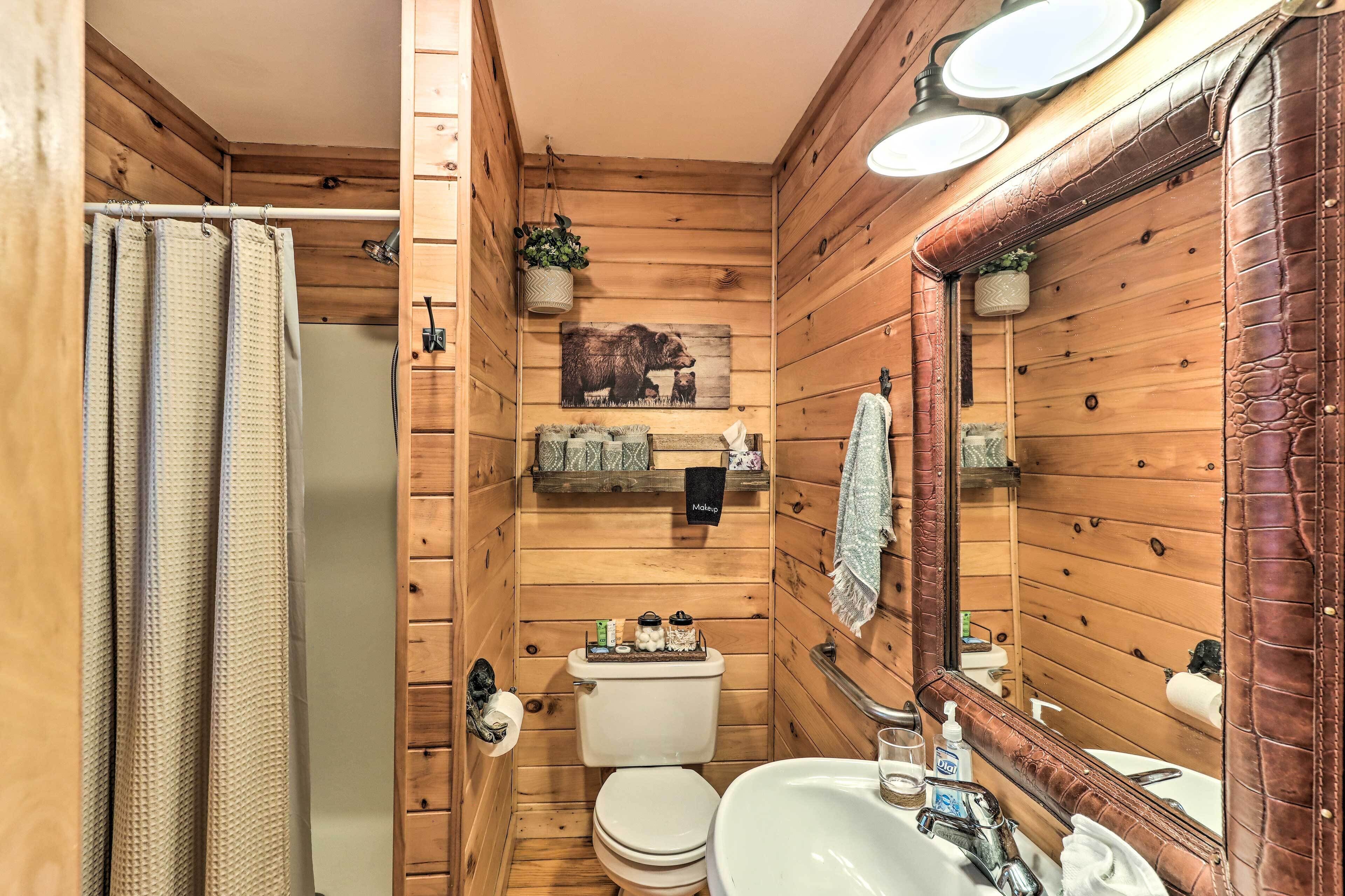 Baño de cabaña de madera con inodoro, lavamanos y ducha. Hay repisas con artículos de aseo y un espejo con una toalla colgada.