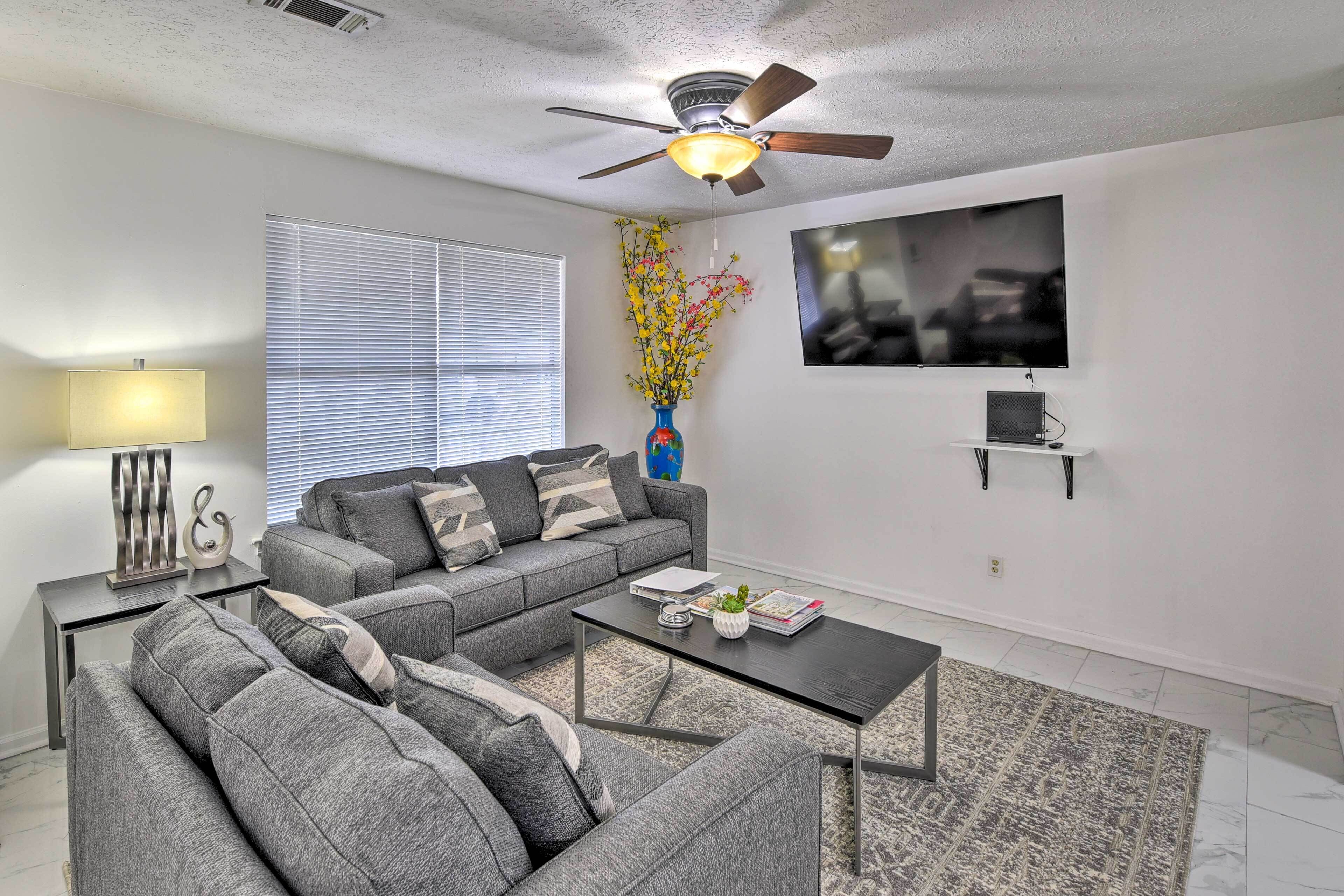 A living room with a grey sectional sofa, a coffee table, a TV mounted on the wall, and a ceiling fan.