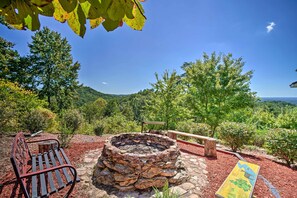 A stone fire pit surrounded by a bench and a picnic table, set against a backdrop of lush greenery and a clear blue sky. - Eclectic Ranger Home w/ Mtn Views + Hot Tub! (Ranger)