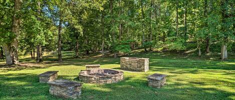 A stone fire pit surrounded by stone benches in a grassy park area with trees.