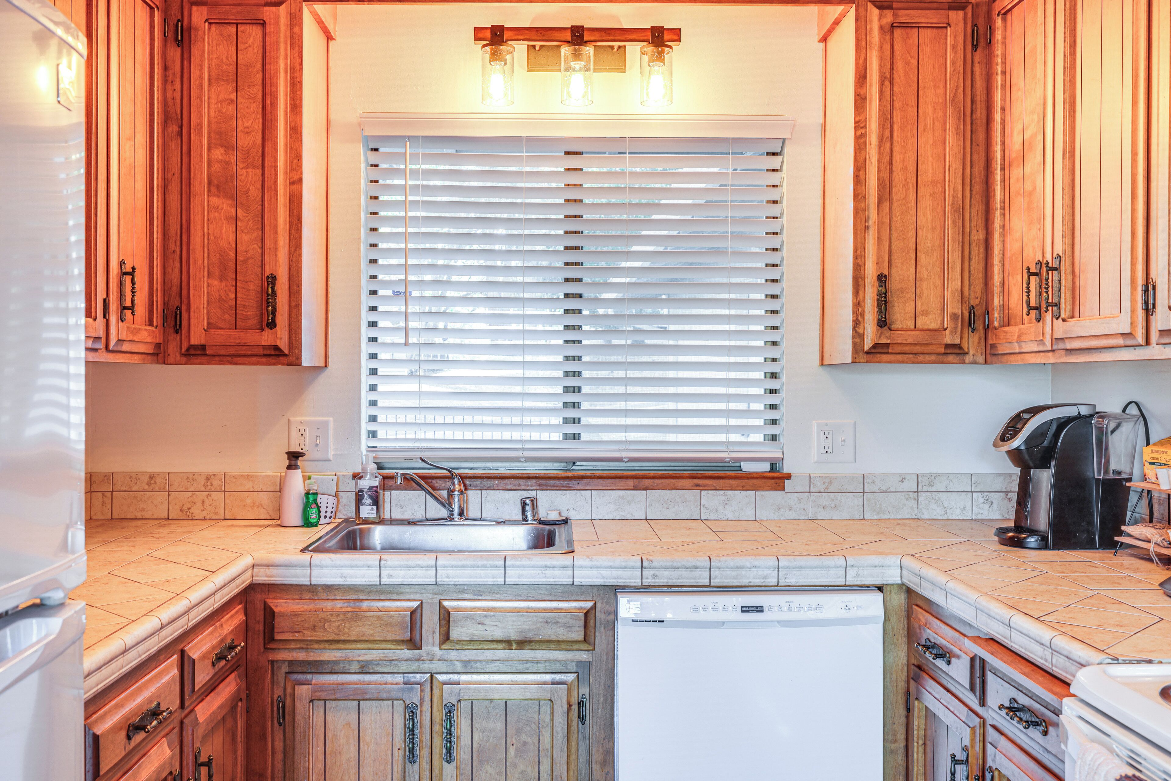 A kitchen with wooden cabinets, a built-in dishwasher, a window with blinds, a coffee maker, and a sink.