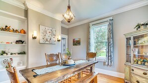Een eetkamer met een houten tafel, bankjes en stoelen. Er is een kroonluchter, een raam met gordijnen en een kast met glazen deuren.