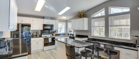 A modern kitchen with white cabinets, a large island with bar stools, and a view of the outdoors through large windows.