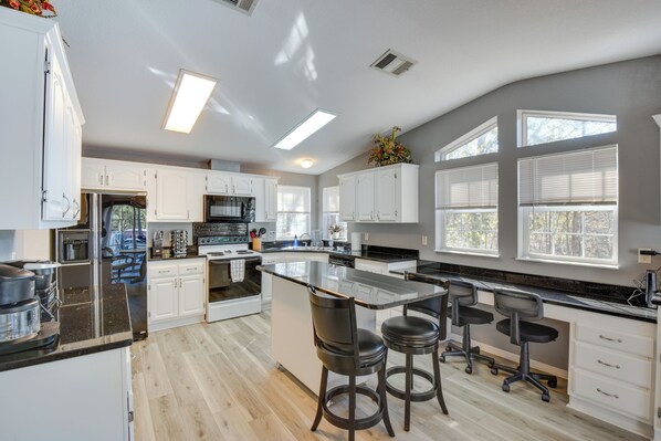 A modern kitchen with white cabinets, a large island with bar stools, and a view of the outdoors through large windows. - Waterfront Lake Hartwell Home w/ Dock & Fire Pit (Hartwell)