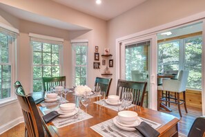 A dining room with a wooden table set for four, surrounded by chairs, with a view of the outdoors through large windows. - Helen Retreat w/ Hot Tub, 2 Mi to Downtown! (Helen)