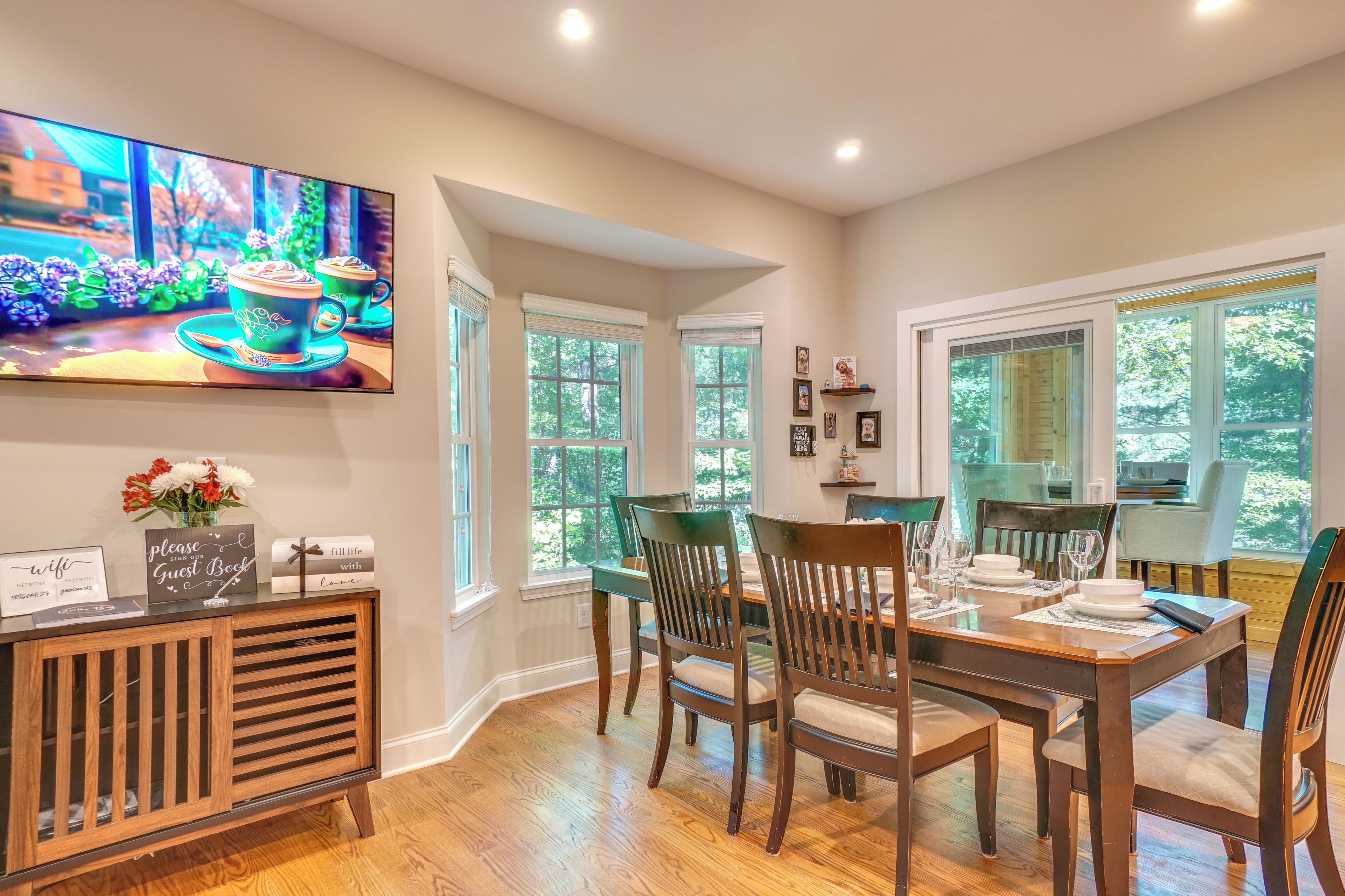 A dining area with a wooden table set for four, chairs, a TV mounted on the wall, and a guest book on the console table.