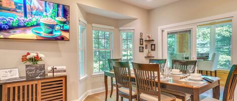 A dining area with a wooden table set for four, chairs, a TV mounted on the wall, and a guest book on the console table.