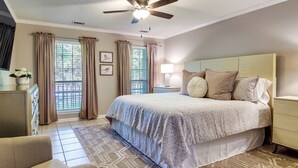 Ein Schlafzimmer mit einem Bett, Deckenventilator, Nachttisch, Lampe, Schminktisch und einem Fenster mit Vorhängen.