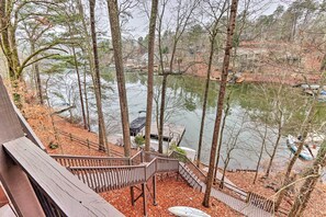 A wooden deck with a view of a lake, surrounded by trees and a few buildings in the background.