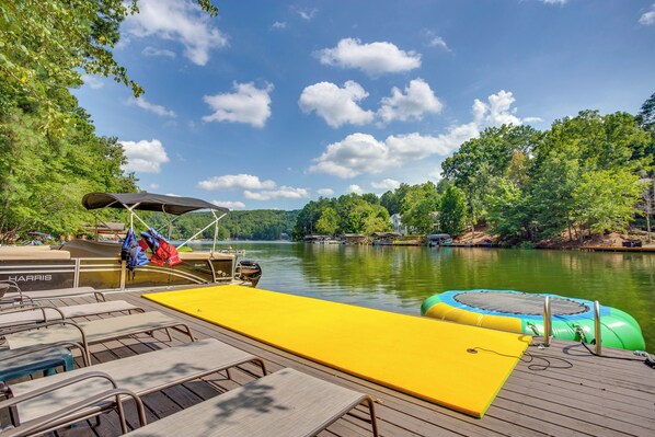 A wooden deck with lounge chairs and a yellow mat, overlooking a calm lake with boats and greenery.