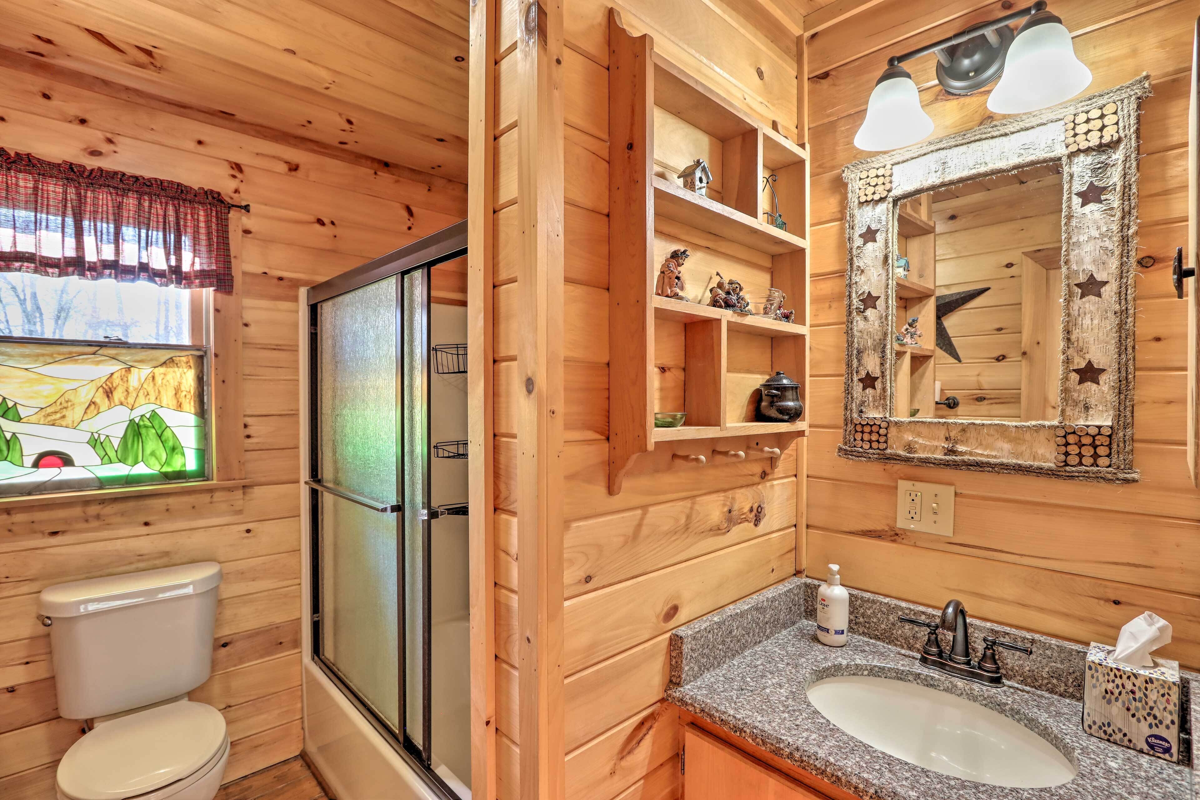 A wooden cabin bathroom with a toilet, shower, vanity with a sink, and shelves with decorative items.