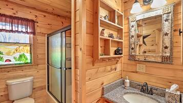 A wooden cabin bathroom with a toilet, shower, vanity with a sink, and shelves with decorative items.