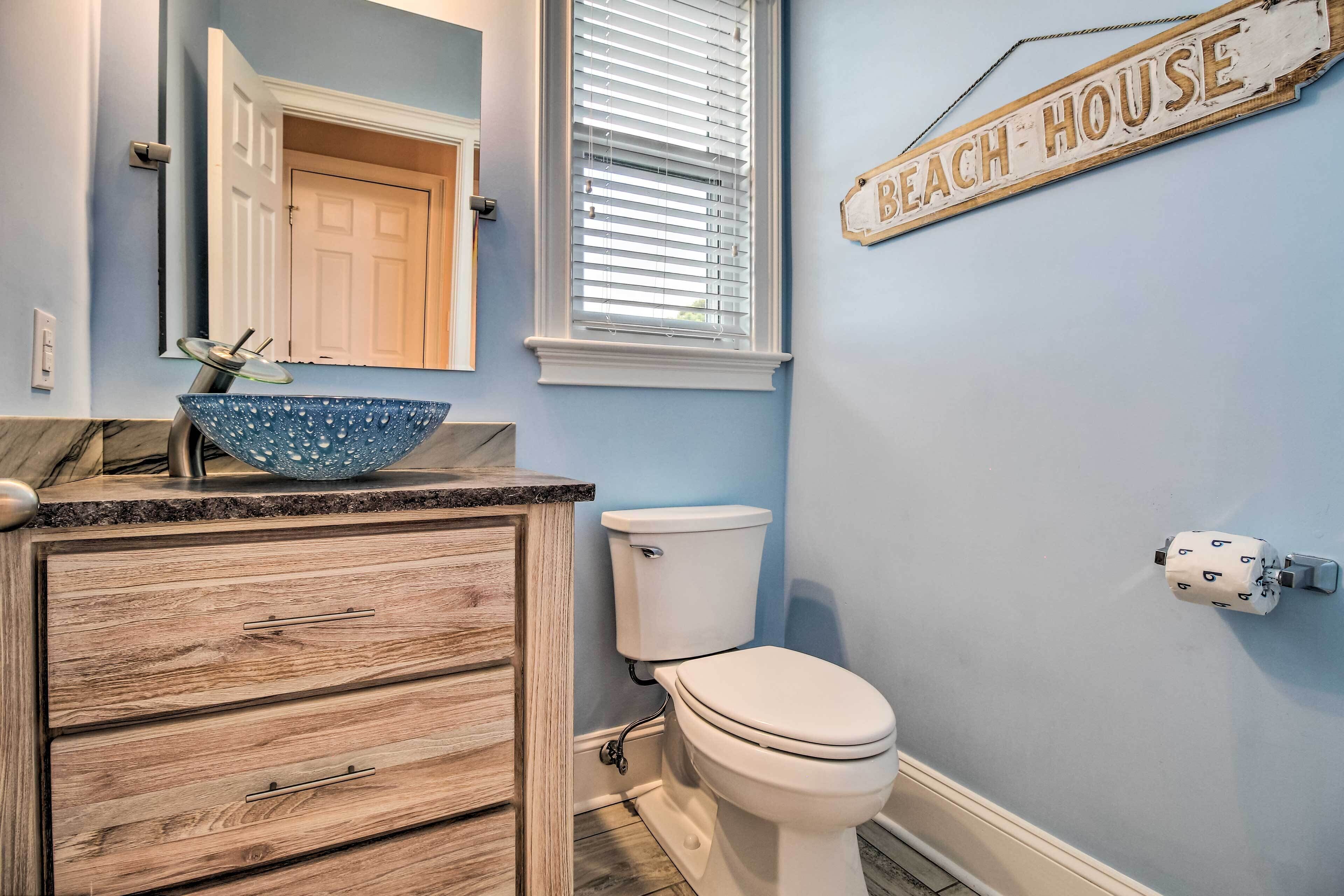 A bathroom with a wooden vanity, a blue bowl sink, a toilet, and a 'Beach House' sign on the wall.