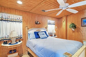 Une chambre à coucher aux murs lambrissés, comprenant un lit, deux tables de chevet, un ventilateur de plafond et une fenêtre avec des rideaux.