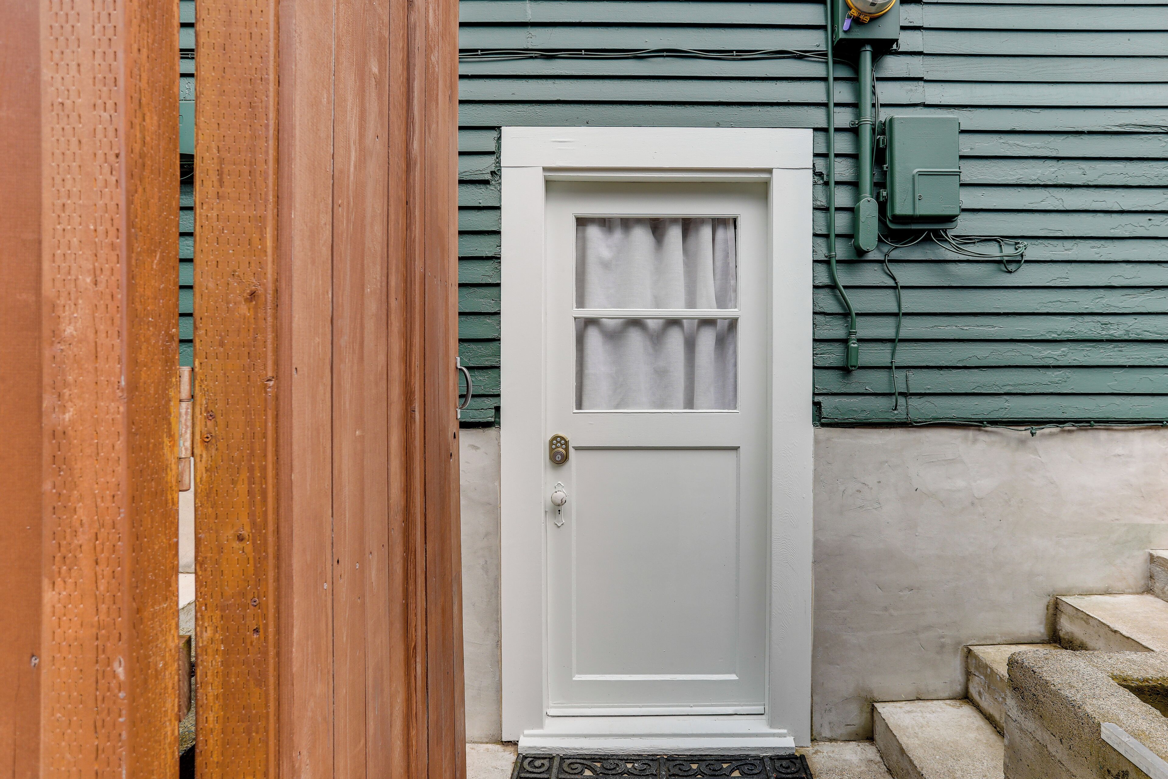 A white door with a small window and a doorknob, set against a green wooden wall with a utility box and wires visible.