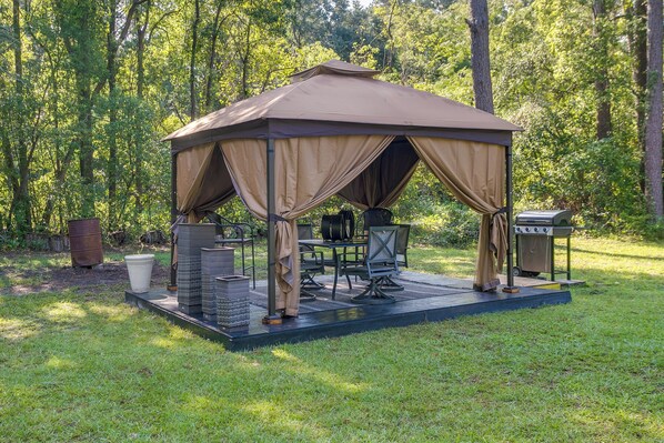 A gazebo with a table and chairs, surrounded by a grassy area and trees.