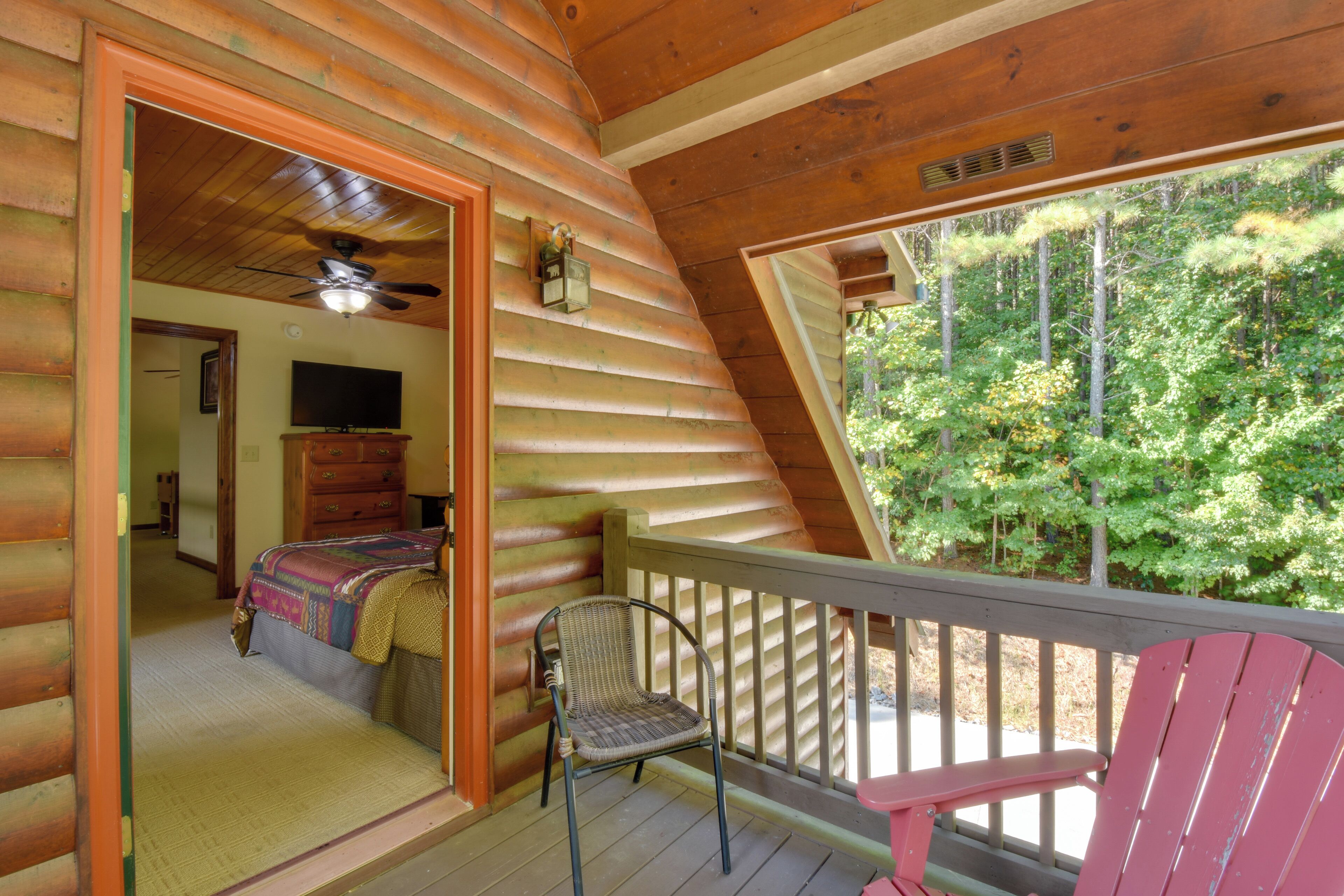 Une cabane comprenant une chambre avec un lit, une télévision et un ventilateur de plafond. La chambre donne sur une terrasse en bois, où se trouvent une chaise et un fauteuil Adirondack rouge.