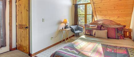 Un dormitorio con techo de madera, una cama con ropa de cama estampada, un escritorio con lámpara y una ventana con cortinas.