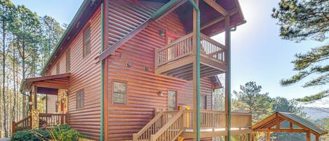 A two-story log cabin with a balcony, a stone foundation, and a small porch area.