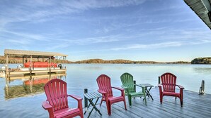 Un muelle de madera con cuatro sillas Adirondack y una mesa, dando vista a aguas tranquilas con un barco amarrado al fondo.