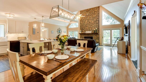 Una cocina amplia con una mesa de comedor de madera dispuesta para seis personas, una chimenea de piedra y ventanas grandes.
