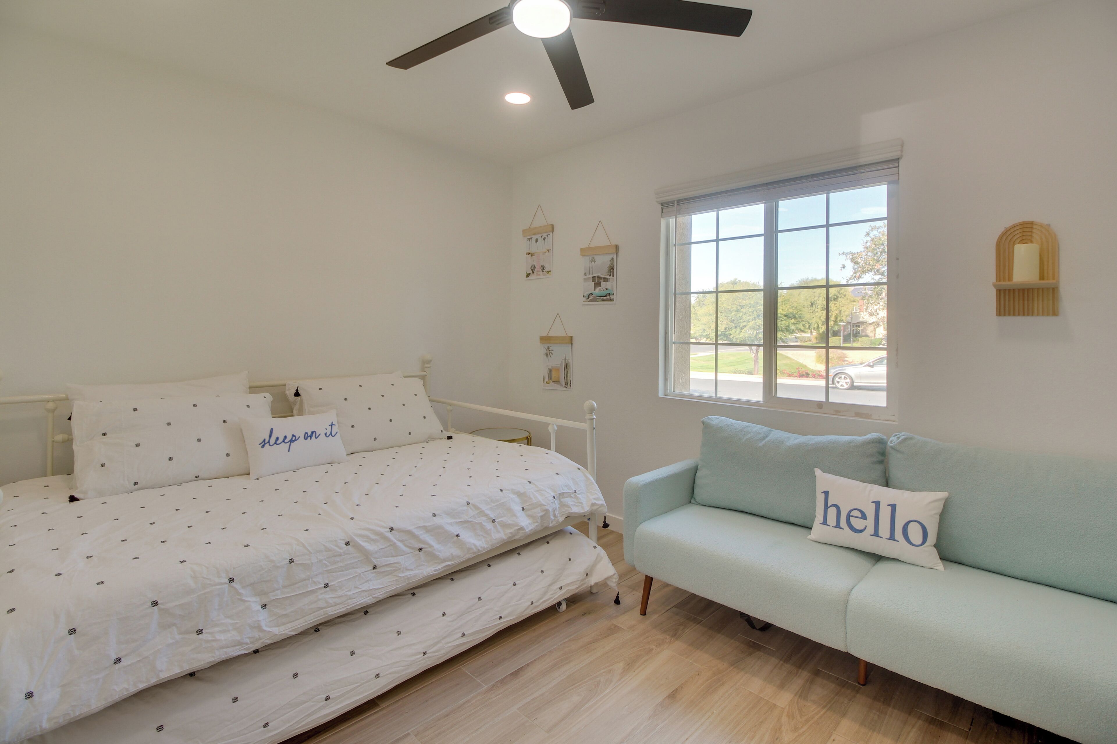 A bedroom with a bed, a light blue sofa, a ceiling fan, a window with a view, and wall-mounted decorations.