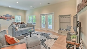 A spacious living room with a grey sofa, a coffee table, and a large floral rug. There are two windows with blinds, a door leading outside, and a staircase with a wooden railing. - Cozy Tennessee Country Retreat on 5 Acres! (Pegram)