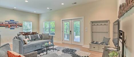 A spacious living room with a grey sofa, a coffee table, and a large floral rug. There are two windows with blinds, a door leading outside, and a staircase with a wooden railing.