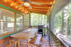 Um deck de madeira com mesa, bancos, ventilador de teto, luzes de cordão e churrasqueira.