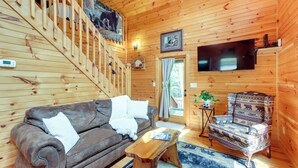 L'intérieur d'un chalet en bois, avec un canapé en cuir marron, un fauteuil à motifs, une table basse en bois, un escalier et un téléviseur fixé au mur.