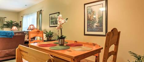 A dining area with a wooden table, chairs, and a vase with flowers. There are framed pictures on the wall and a lamp on the table.
