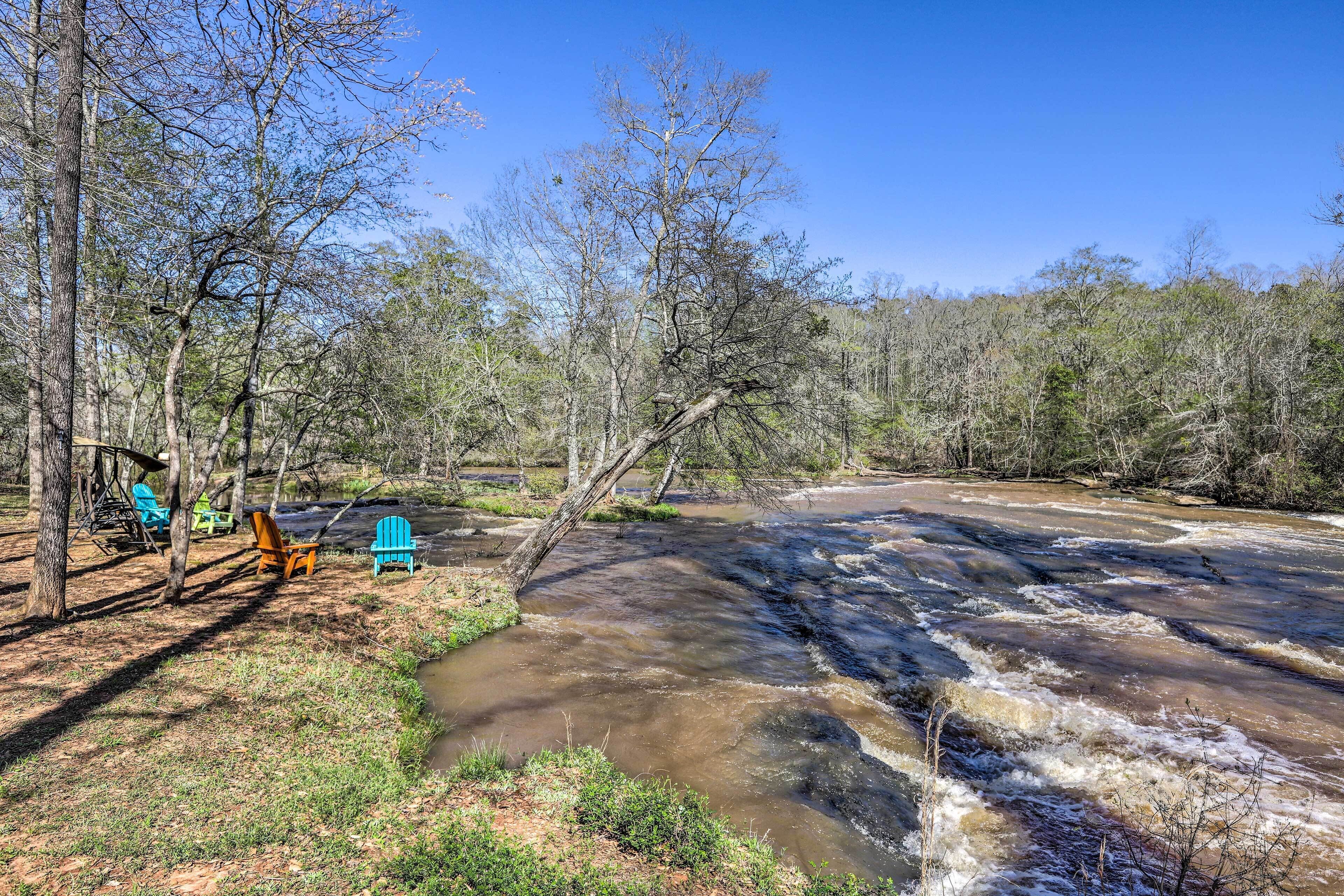 A river with a wooden bridge, surrounded by trees and outdoor seating.