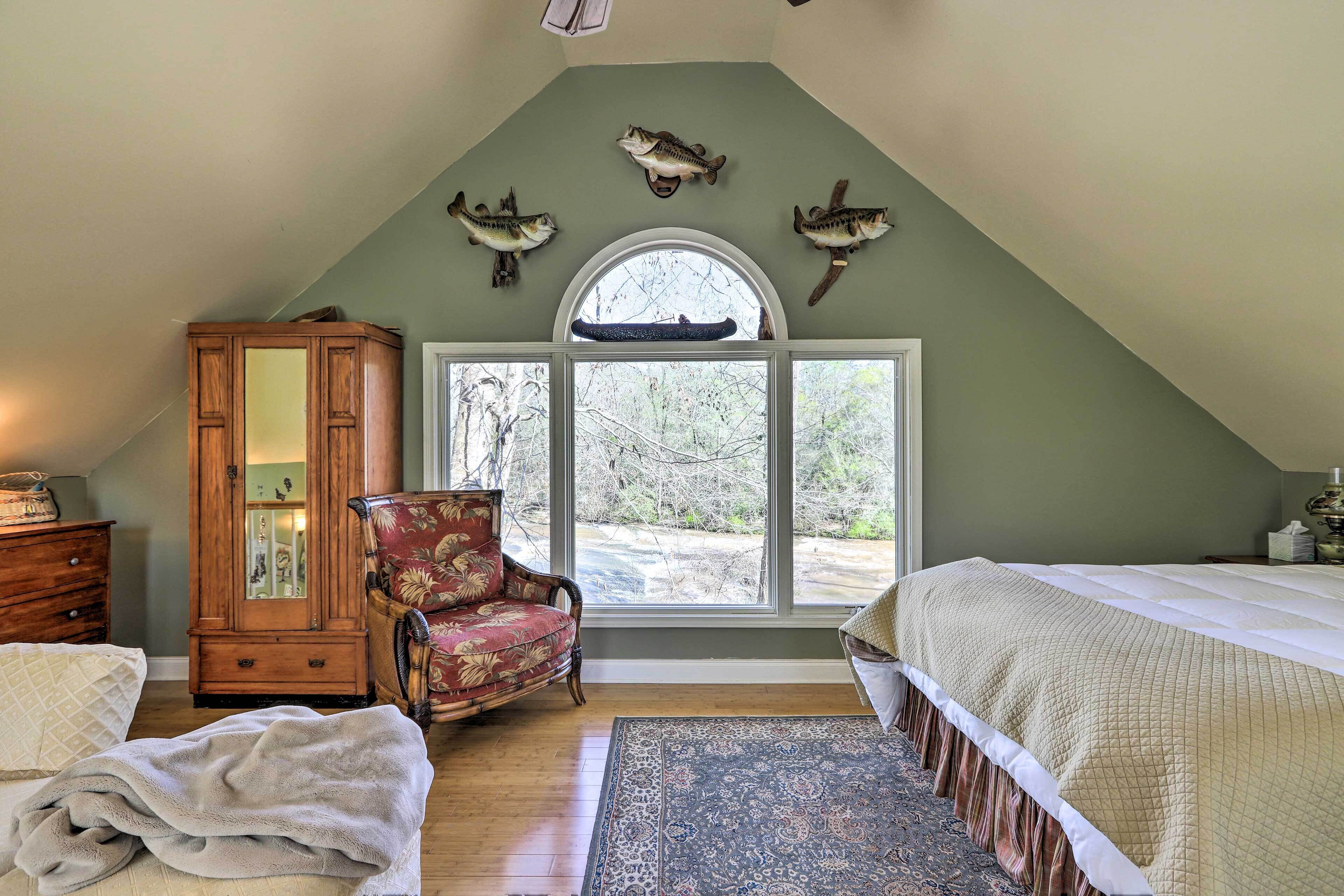 A bedroom with a sloped ceiling, a large window, a wooden cabinet, a floral armchair, a bed with a quilted bedspread, and a patterned rug.